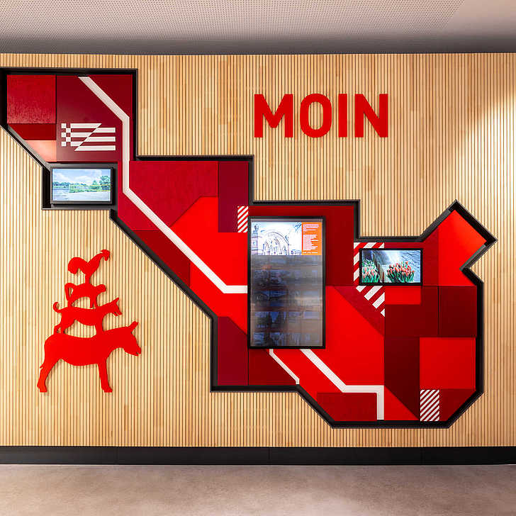 Eine holzvertäfelte Wand. Oben steht in rot der große Schriftzug "MOIN". Darunter befinden sich mehrere Bildschirme und eine rote Silhouette der Bremer Stadtmusikanten.