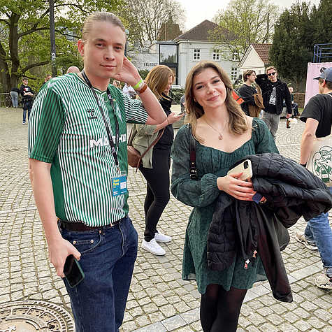 Benedict und Alea aus dem Team des Filmfests Bremen stehen auf dem Vorplatz des Bremer Theaters. Sie lächeln in die Kamera, im Hintergrund sind weitere Gäste und das Festivalzentrum zu sehen.