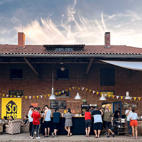 Außenansicht eines Gebäudes mit Ziegelfassade, vor dem sich eine Bar mit mehreren Gästen befindet. Gelbe Wimpelketten und ein Banner mit der Aufschrift '30' schmücken den Bereich, während der Himmel mit leichten Wolken den Hintergrund bildet.