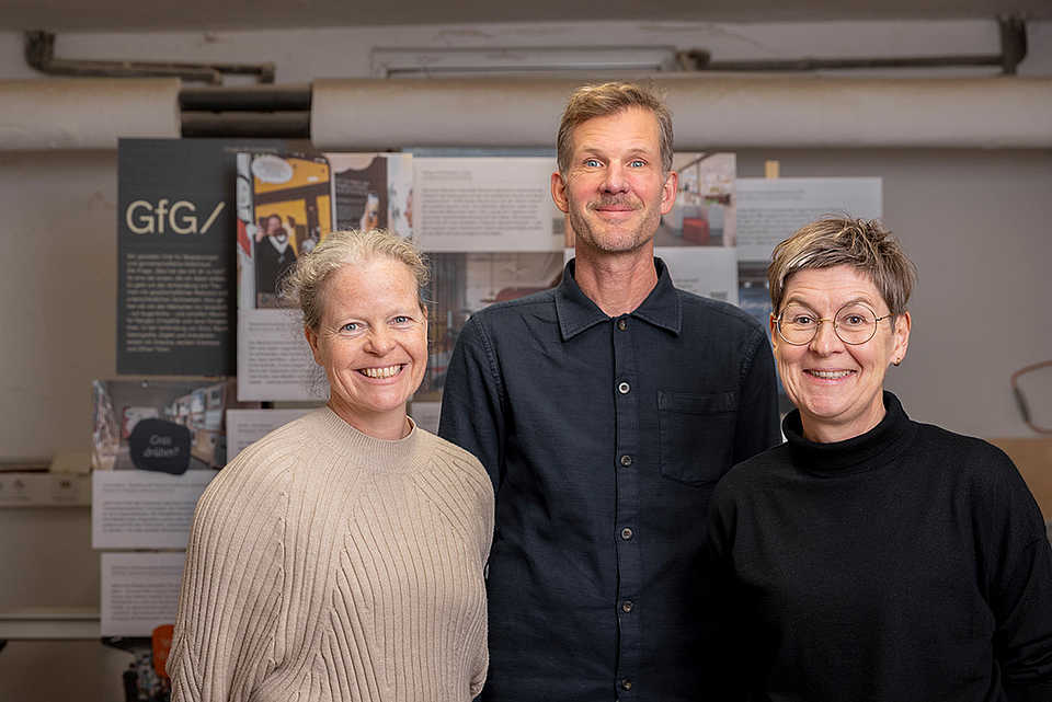 Siena, Carsten und Ulrike aus dem GfG-Team stehen lächelnd vor einer Präsentationswand mit Ausstellungsprojekten.