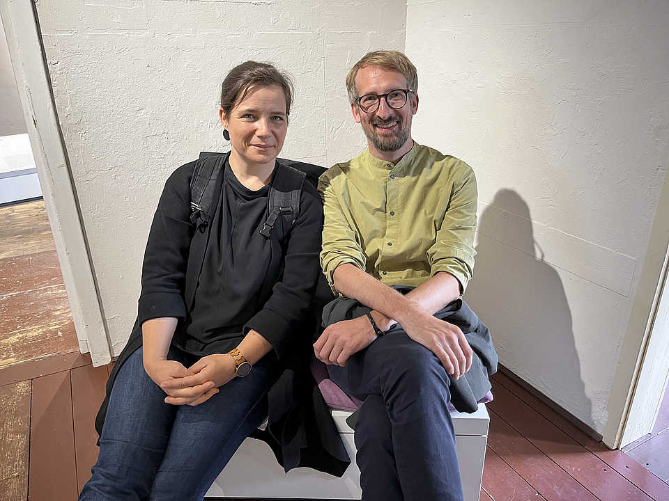 Eine Frau und ein Mann sitzen nebeneinander auf einer Bank in einem Ausstellungsraum. 