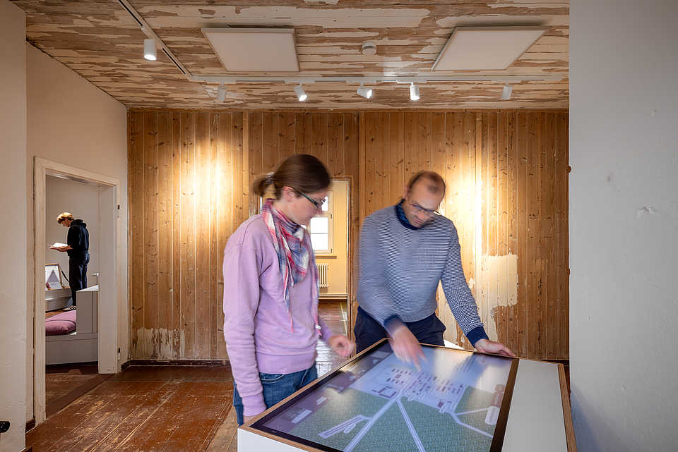 Zwei Personen stehen vor einem interaktiven Display in einem Raum mit Holzvertäfelung. Eine Person deutet auf den Bildschirm, während die andere zuhört.