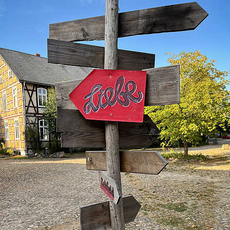 Ein Holzwegweiser mit mehreren Richtungsanzeigen, bei dem ein rotes Schild mit dem Wort "Liebe" im Vordergrund hervorsticht. Im Hintergrund ist ein Fachwerkhaus und eine sonnige, grüne Umgebung zu sehen.