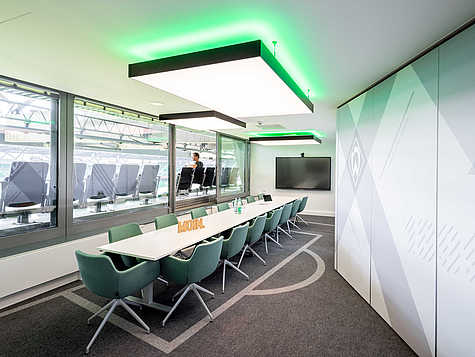 Ein moderner Konferenzraum mit Blick auf ein Stadion. Der Raum ist mit einem langen weißen Tisch und grünen Stühlen ausgestattet. An der Decke befinden sich große rechteckige Lampen mit grünem Licht. Eine Wand zeigt das Werder Bremen Logo. Vor den Fenstern sind Sitzreihen des Stadions zu sehen, wo eine Person sitzt. Der Raum ist hell und bietet eine inspirierende Aussicht auf das Stadion.