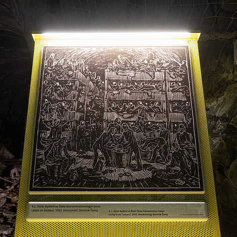 Beleuchtete Ausstellungstafel mit einem Holzschnitt von Dominik Černý aus dem Jahr 1953, der das Leben von Häftlingen im im Stollen darstellt.“