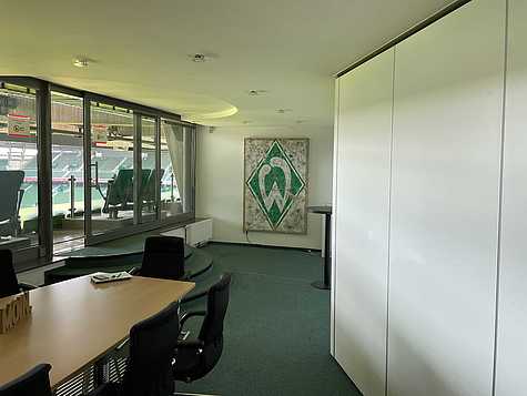 Ein Büroflur mit grünen Wänden und Teppichboden. An der rechten Wand befindet sich das Logo von Werder Bremen und der Schriftzug 'LEBENSLANG GRÜN-WEISS.' Links sind mehrere Türen und ein Feuerlöscher zu sehen. Am Ende des Flurs stehen ein Drucker und ein Tisch, daneben führt eine kleine Treppe zu einem anderen Bereich.