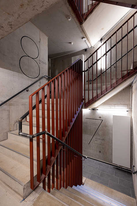 Treppenhaus mit roten Geländern und Betonwänden. An der Wand sind Stockwerksnummern '7' und '8' stilvoll gestaltet angebracht.