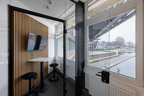 Kleine Glaskabine mit Holzlamellenwand, zwei schwarzen Hockern und einem weißen Stehtisch. An der Wand hängt ein Bildschirm. Große Fenster geben den Blick nach draußen frei.