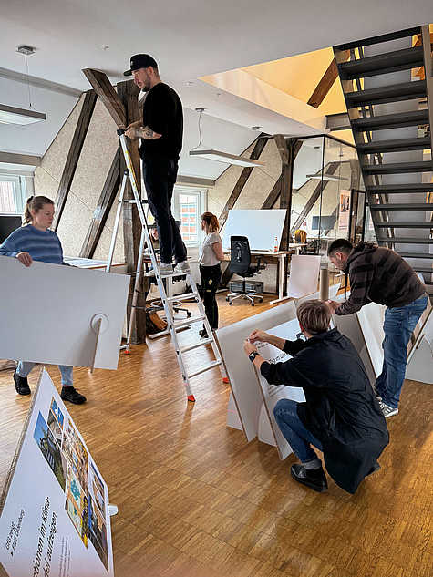 Team beim Aufbau einer Ausstellung in einem modernen Raum mit sichtbaren Holzbalken und einer Metalltreppe. Eine Person steht auf einer Leiter, während andere Plakate und Ausstellungstafeln vorbereiten.
