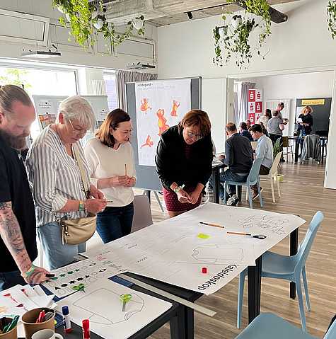 Kleine Arbeitsgruppe entwickelt Ideen an einem Stehtisch und diskutiert gemeinsam über Entwürfe und Workshopmaterialien.