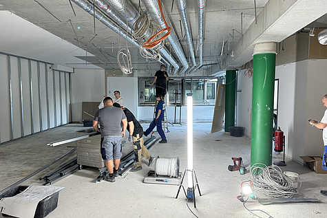 Mehrere Handwerker arbeiten in einem großen, unfertigen Büroraum. Kabel hängen von der Decke, Rohre und Lüftungsschächte sind sichtbar. In der Mitte steht eine helle Baustellenleuchte, auf dem Boden liegen Werkzeuge, Kabelrollen und Baumaterialien. Im Hintergrund steht ein Gerüst unter einer abgehängten Decke.