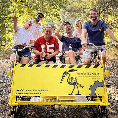 Gruppe von sechs Personen, die auf einer gelben Fahrraddraisine der WendenTEC GmbH sitzen und fröhlich in die Kamera lachen. Im Hintergrund ist eine bewaldete Landschaft zu sehen.