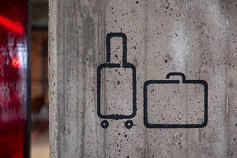 Eine minimalistische, auf Beton gemalte Piktogramm-Darstellung von einem Rollkoffer und einem Aktenkoffer, platziert auf einer grob strukturierten Betonwand.