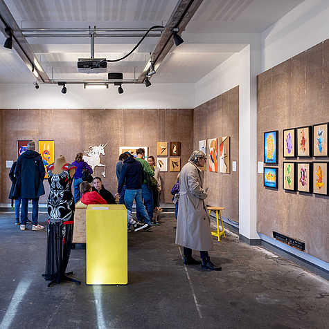 Ein heller Ausstellungsraum mit Besucher:innen, die Kunstwerke an den Wänden betrachten, darunter Gemälde und eine Einhorn-Skulptur. Im Vordergrund steht eine bekleidete Schaufensterpuppe neben einem gelben Podest.