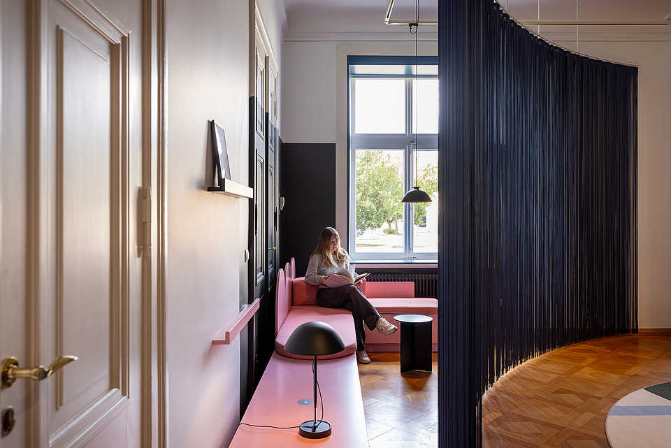 Eine Person sitzt auf einer rosa Couch und liest ein Buch. Die Couch steht an einer Wand mit schwarzen Türen, und durch das große Fenster im Hintergrund fällt natürliches Licht in den Raum. Vor der Couch steht ein kleiner schwarzer Tisch, und eine schwarze Lampe beleuchtet die Szene.