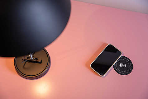 Ein schwarzes, modernes Tischlampenfuß steht auf einer rosa Oberfläche. Daneben liegt ein Smartphone auf einem kabellosen Ladepad, das mit einem Batteriesymbol gekennzeichnet ist. Das Smartphone befindet sich leicht schräg auf dem Ladepad, und die Oberfläche reflektiert sanft das Licht der Lampe.