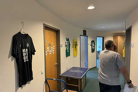 Flur in einem Bürogebäude mit mehreren an der Wand hängenden Fußballtrikots. An einer Tür kleben Sprüche und Vereinsaufkleber, davor steht eine kleine Tischtennisplatte. Eine Person mit grauem T-Shirt geht durch den Flur .