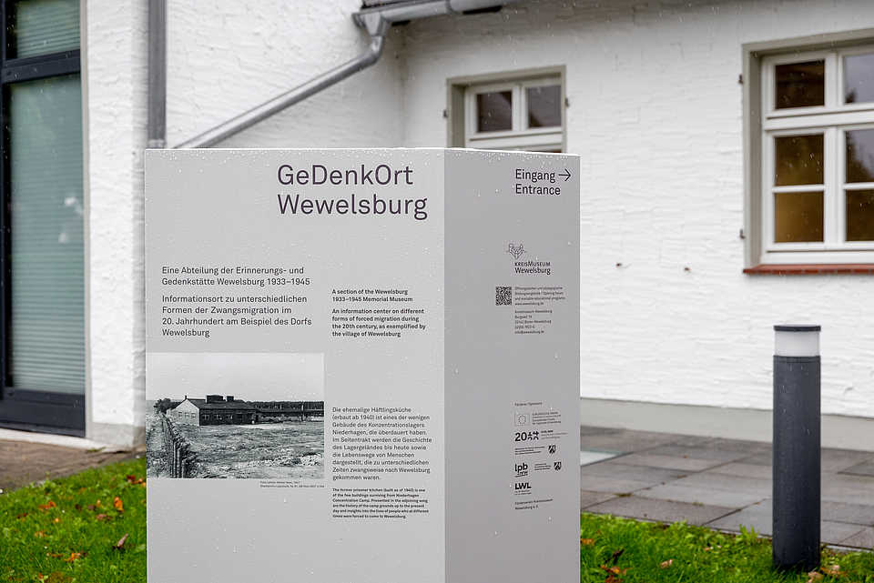 Eine große Stele mit der Aufschrift „GeDenkOrt Wewelsburg“ und Informationen in Deutsch und Englisch. Im Hintergrund ist die Fassade eines weißen Gebäudes mit Fenstern und einer Regenrinne zu sehen.