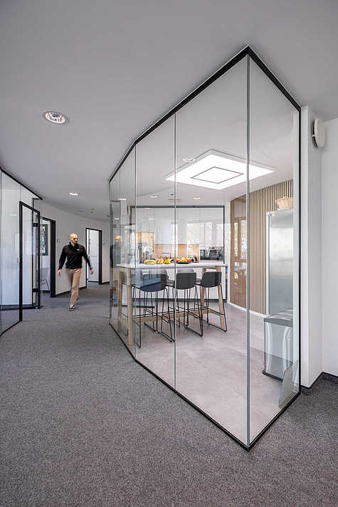 Offener Büroflur mit gebogenen Glaswänden. Rechts befindet sich eine moderne Küche mit Tresen und Barhockern, dahinter ein Mann im schwarzen Pullover, der durch den Flur geht.