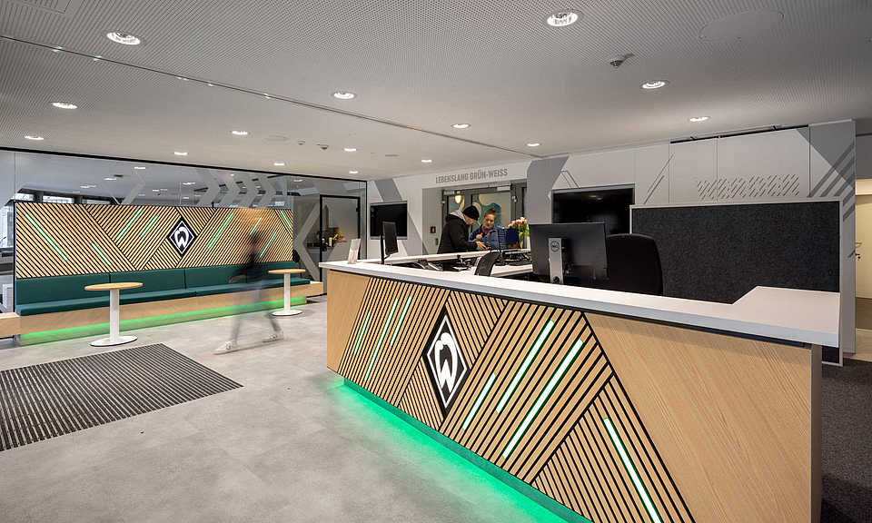Empfangsbereich mit modernem Tresen und Werder-Branding in Holzoptik mit grünen Lichtelementen. Links im Bild eine Sitzbank mit grünem Polster und ebenfalls beleuchteter Holzverkleidung. Zwei Personen stehen am Empfang, im Hintergrund der Schriftzug ‚Lebenslang grün-weiß‘.