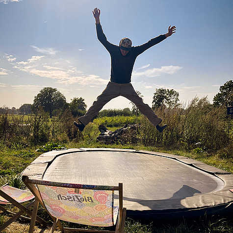 Person springt mit ausgestreckten Armen und Beinen auf einem Trampolin in einer ländlichen Umgebung. Im Vordergrund steht ein bunter Liegestuhl.