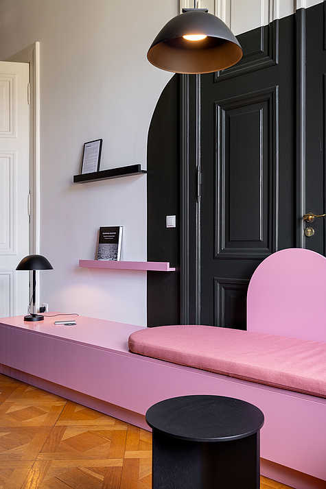 Ein moderner Sitzbereich mit einer rosa Bank, die vor einer großen schwarzen Tür steht. Über der Bank befinden sich zwei minimalistische Regale, auf denen ein Buch und ein gerahmter Text stehen. Eine schwarze Lampe hängt von der Decke und beleuchtet den Bereich. Ein kleiner, runder schwarzer Tisch steht vor der Bank, und auf der rosa Ablagefläche neben der Bank liegt ein Smartphone.