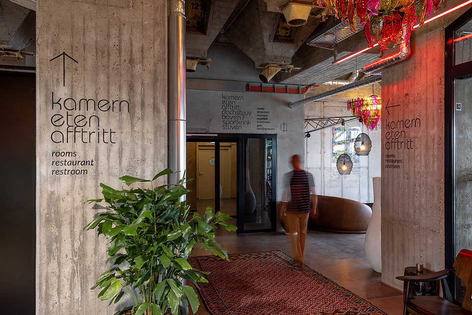 Eine moderne Betonwand in einem Gebäude, mit einer Beschilderung, die "kamern eten afftritt" (rooms, restaurant, restroom) in schlichten schwarzen Buchstaben anzeigt. Im Vordergrund steht eine große grüne Pflanze, während im Hintergrund eine Person vorbeigeht.