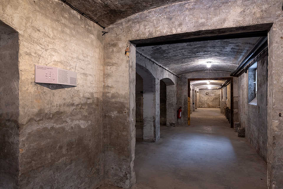 Ein langer, gewölbter Kellerflur aus Stein mit mehreren Durchgängen und einer Wandtafel auf der linken Seite. Im Hintergrund sind weitere Räume zu erkennen.