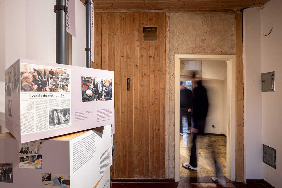 Ausstellungselement mit Bildern und Texten an einer Holzwand. Zwei Personen bewegen sich im Hintergrund durch eine offene Tür.