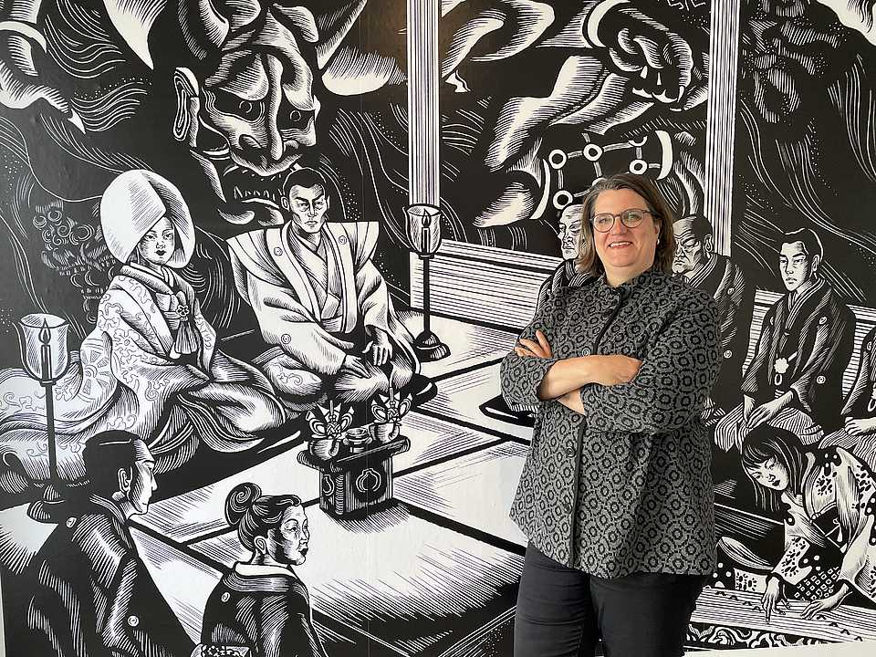 Eine Frau mit kurzen Haaren und Brille steht mit verschränkten Armen vor einer großen Wandillustration. Die Illustration zeigt eine Szene im traditionellen japanischen Stil, mit mehreren Figuren in kimonos, darunter eine Frau mit einem großen Kopfschmuck und ein Mann in formeller Kleidung. Im Hintergrund ist ein großes, dämonisches Gesicht zu sehen. Die Frau trägt eine graue Jacke mit einem geometrischen Muster und lächelt in die Kamera.
