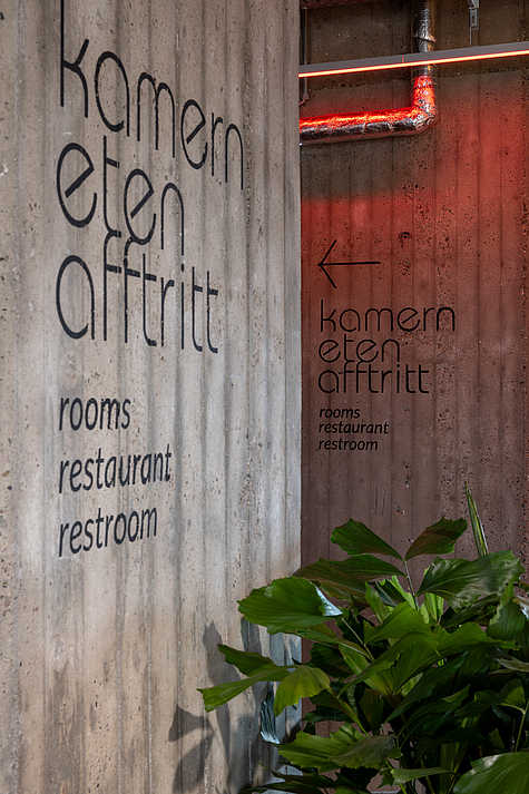 Eine Betonwand mit der Beschilderung "kamern eten afftritt" (rooms, restaurant, restroom), die mit einem roten Licht leicht angestrahlt wird. Neben der Wand steht eine große Pflanze.