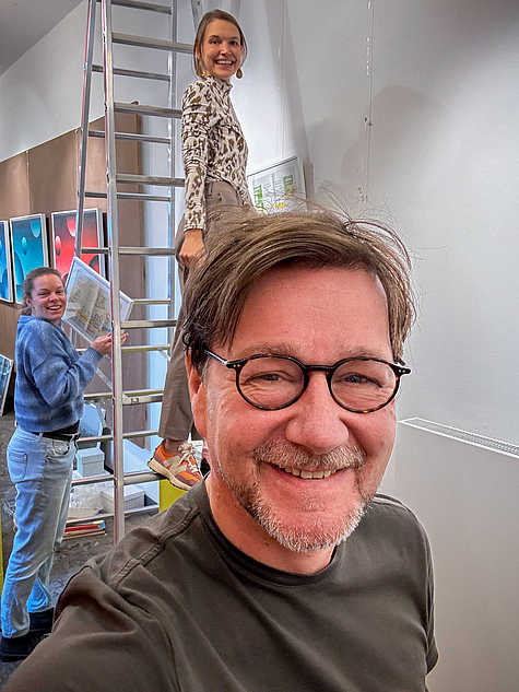 Drei Personen beim fröhlichen Aufbau einer Ausstellung: Im Vordergrund ein Mann mit Brille, im Hintergrund zwei Frauen, eine davon steht auf einer Leiter.