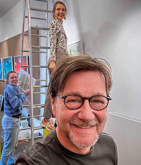 Drei Personen beim fröhlichen Aufbau einer Ausstellung: Im Vordergrund ein Mann mit Brille, im Hintergrund zwei Frauen, eine davon steht auf einer Leiter.