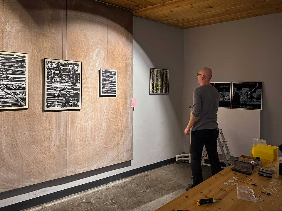 Ein Mann hängt Schwarz-Weiß-Kunstwerke mit abstrakten Mustern an eine Ausstellungswand. Ein Holztisch im Vordergrund ist mit Werkzeugen und Materialien bedeckt.