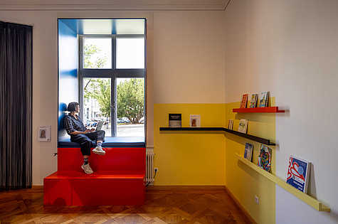 Eine Person sitzt in einer gemütlichen Fensterbank mit einem Laptop auf dem Schoß. Die Fensterbank ist in Blau und Rot gehalten und befindet sich in einem modern gestalteten Raum mit gelben Wänden. Rechts im Bild befinden sich farbige Wandregale mit verschiedenen Büchern. Durch das große Fenster im Hintergrund ist eine parkähnliche Landschaft mit Bäumen zu sehen, die den Raum mit Tageslicht erhellt.