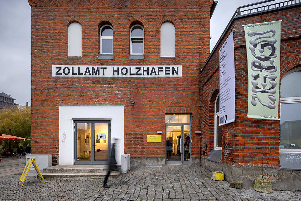 Fassade des historischen Zollamts Holzhafen mit Bannern, die die Ausstellung 'GfG Zeigen' ankündigen. Menschen gehen vorbei, während im Inneren Kunstwerke durch beleuchtete Fenster sichtbar sind.