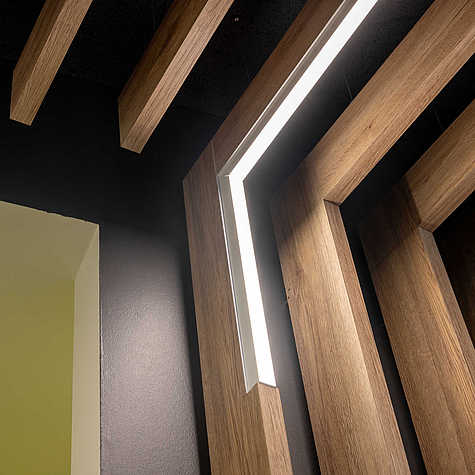 Detailansicht einer Decke mit vertikalen Holzlamellen und integrierter L-förmiger LED-Leuchte, vor dunkler Wand.