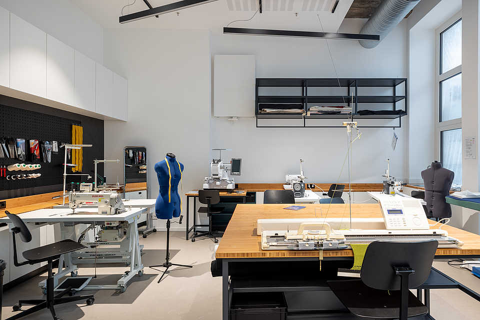 Eine Werkstatt mit Nähmaschinen, Schneidertisch und Schneiderpuppe. Die Einrichtung ist modern und funktional, mit Materialien und Werkzeugen an einer schwarzen Lochwand organisiert.