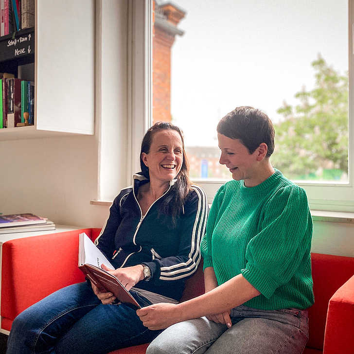 Autorin Sabine Doff und die Redakteurin Ninja Hofmann sitzen gemeinsam auf einem roten Sofa vor einem Fenster und blättern durch das Buch "Das Klassentreffen".