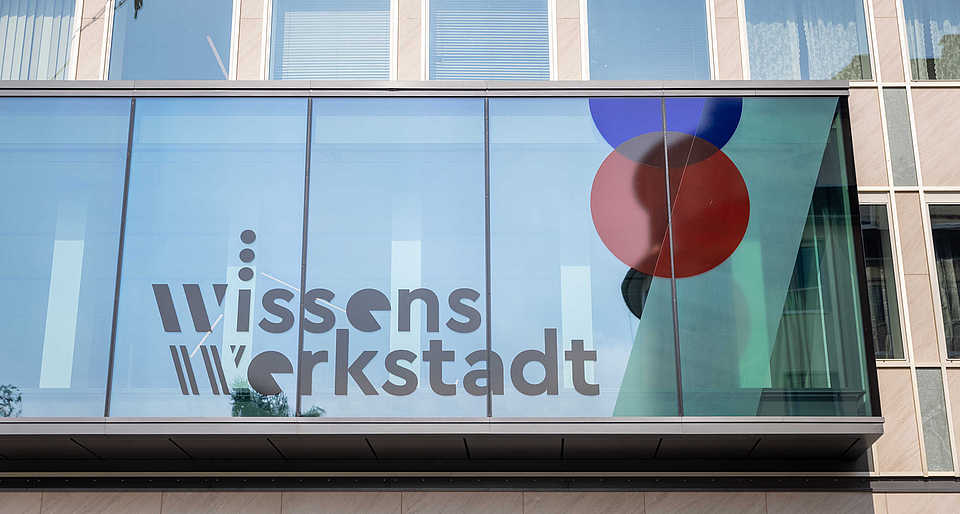 Ein modernes Gebäude mit einer Glasfassade, die das Logo der Wissenswerkstadt trägt. Zwei große, farbige Kreise sind dekorativ in die Scheiben integriert.