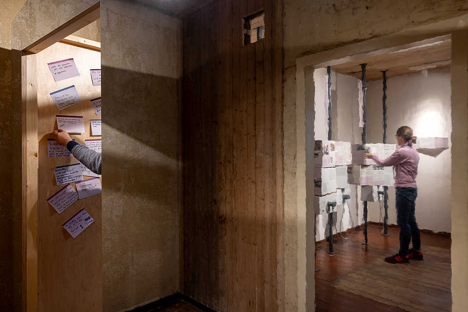 Eine Wand aus Holz mit handgeschriebenen Notizen auf Papierkarten, die an die Wand geheftet sind. Im Hintergrund ist eine Person zu sehen, die an der Wand arbeitet.