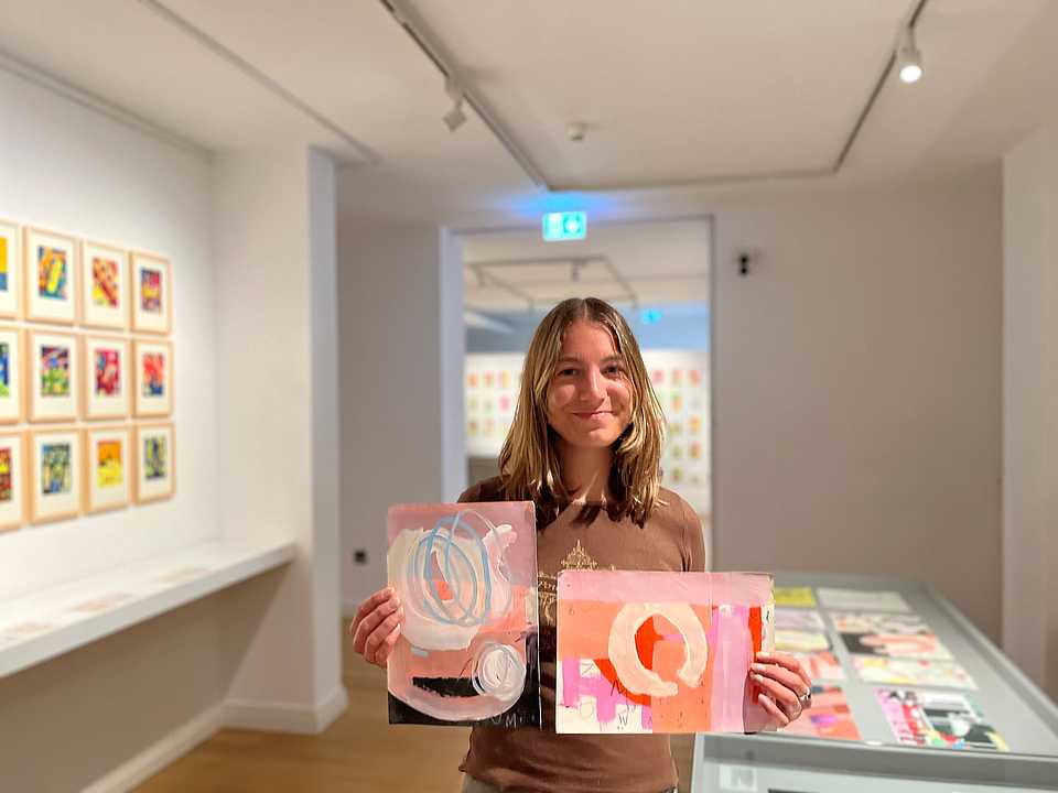 Eine junge Frau mit schulterlangem Haar steht in einer Galerie und hält zwei abstrakte Gemälde in den Händen. Sie lächelt in die Kamera. Im Hintergrund sind weitere Kunstwerke an den Wänden sowie ein großer Tisch mit mehr Kunstwerken unter Glas zu sehen. Die Galerie hat weiße Wände und eine ruhige, gut beleuchtete Atmosphäre.