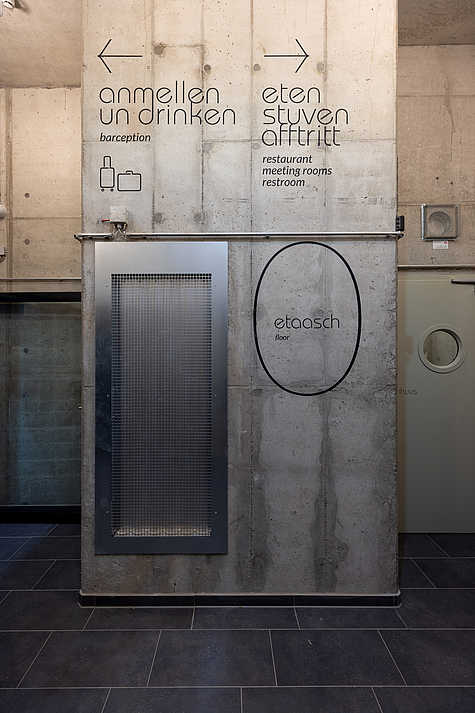 Eine graue Betonwand mit einer Beschriftung in moderner Typografie. Links zeigt ein Pfeil zur „Barception“ und rechts zur „Eten Stuven“. Ein ovaler schwarzer Rahmen hebt das Wort „etaasch floor“ hervor. Darunter befindet sich ein Metallgitter-Element.