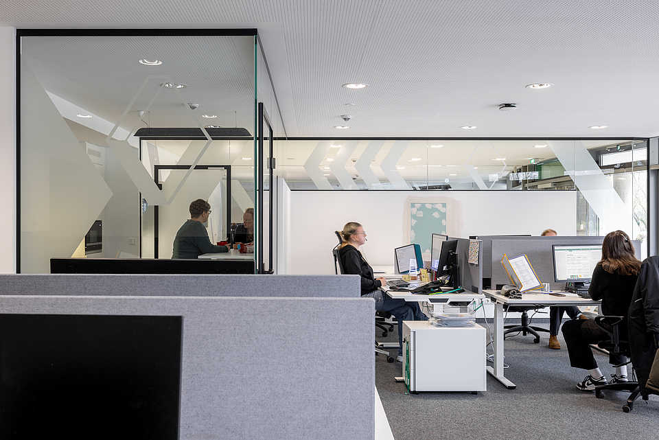Großraumbüro mit mehreren modernen Arbeitsplätzen. Im Vordergrund arbeiten Personen an höhenverstellbaren Schreibtischen. Im Hintergrund ein Glaskubus mit zwei Personen im Gespräch.