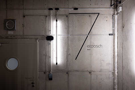Ein grauer Betonwandbereich mit der Aufschrift „7 etaasch floor“ in moderner Typografie. Daneben eine schmale, vertikale Neonröhre, die den Bereich beleuchtet. Eine Tür mit einem runden Fenster ist ebenfalls sichtbar.