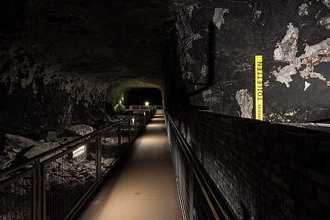 Tunnelgang mit beleuchtetem Steg in der KZ-Gedenkstätte Mittelbau-Dora. Rechts ein gelber Pfeiler mit Aufschrift ‚Toiletten 1944/45‘.