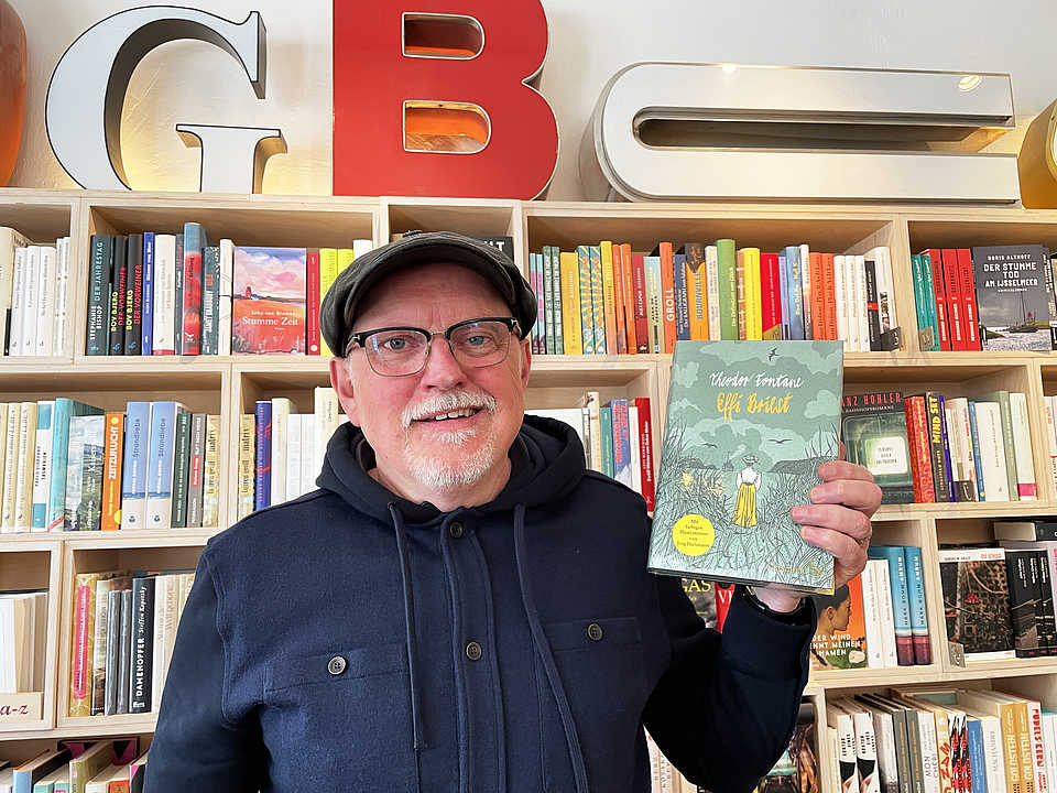 Ein Mann mit Brille und Mütze steht vor einem Bücherregal und hält ein Buch mit dem Titel 'Effi Briest' von Theodor Fontane in der Hand. Er lächelt in die Kamera. Im Hintergrund sind zahlreiche bunte Bücher zu sehen.
