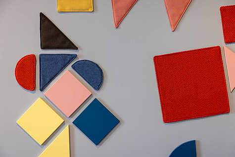 Eine Nahaufnahme von farbigen geometrischen Formen, die an einer Wand angebracht sind. Zu den Formen gehören Halbkreise, Dreiecke, Rechtecke und Quadrate in den Farben Rot, Blau, Gelb, Rosa und Schwarz. Die Texturen der Formen variieren und scheinen teils aus Stoff zu bestehen, was dem Design eine haptische Dimension verleiht.