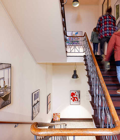 Treppenhaus einer Ausstellung mit aufgehängten Kunstwerken an den Wänden. Mehrere Personen steigen die Treppe hinauf, während verschiedene Kunstwerke die Wände dekorieren.