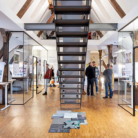 Das Bild zeigt einen modernen Ausstellungsraum mit hohen Decken und sichtbaren Holzbalken. Im Mittelpunkt steht eine freitragende Metalltreppe, die nach oben führt. Links und rechts befinden sich gläserne Räume mit Ausstellungstafeln. Einige Menschen unterhalten sich im Hintergrund, während der Fokus auf der offenen und gut beleuchteten Architektur des Raums liegt. Auf dem Boden sind verschiedene Materialien ausgelegt, möglicherweise Teil der Ausstellung.